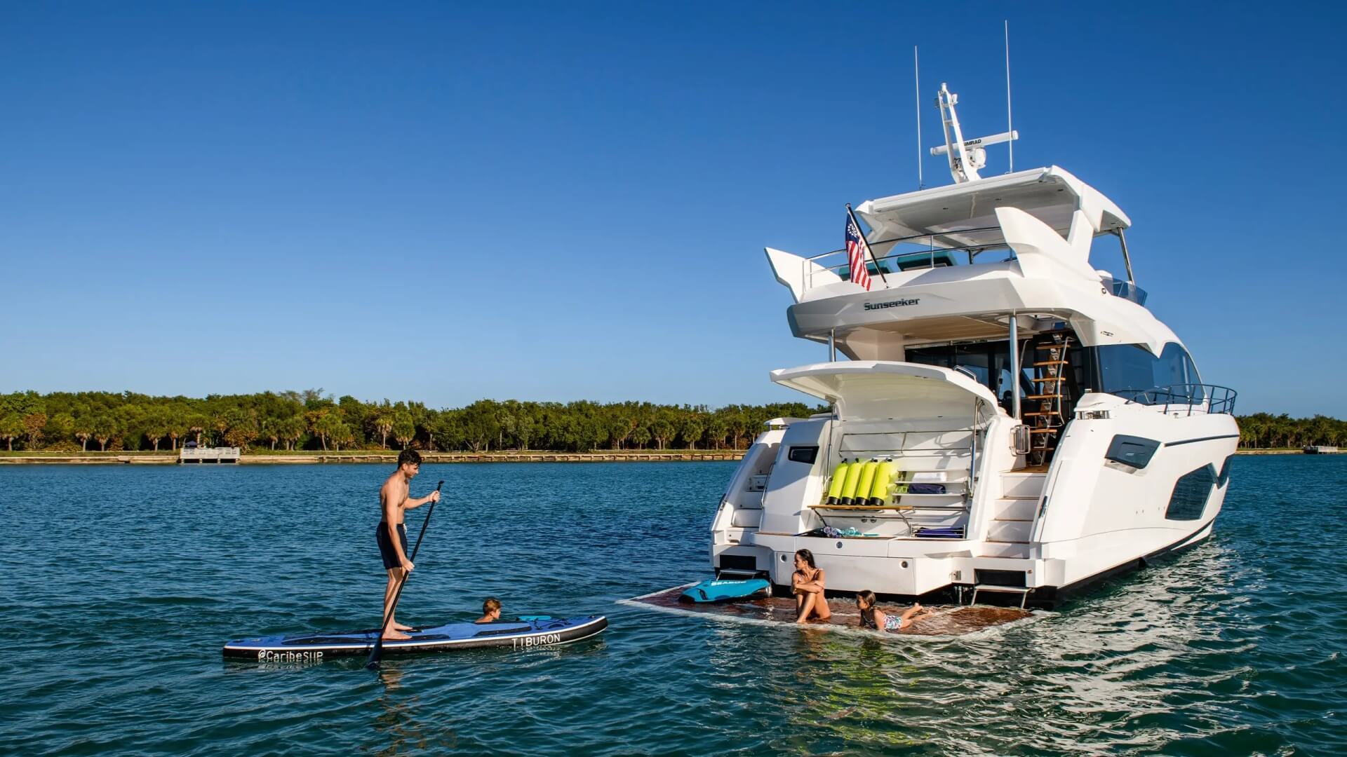 Stern of Sunseeker Manhattan 68 M/Y Apollo yacht - family enjoying their time on a sunny day. Father is on a SUP board, son is swimming, and mother and daughter are sunbaiding on a swim platform